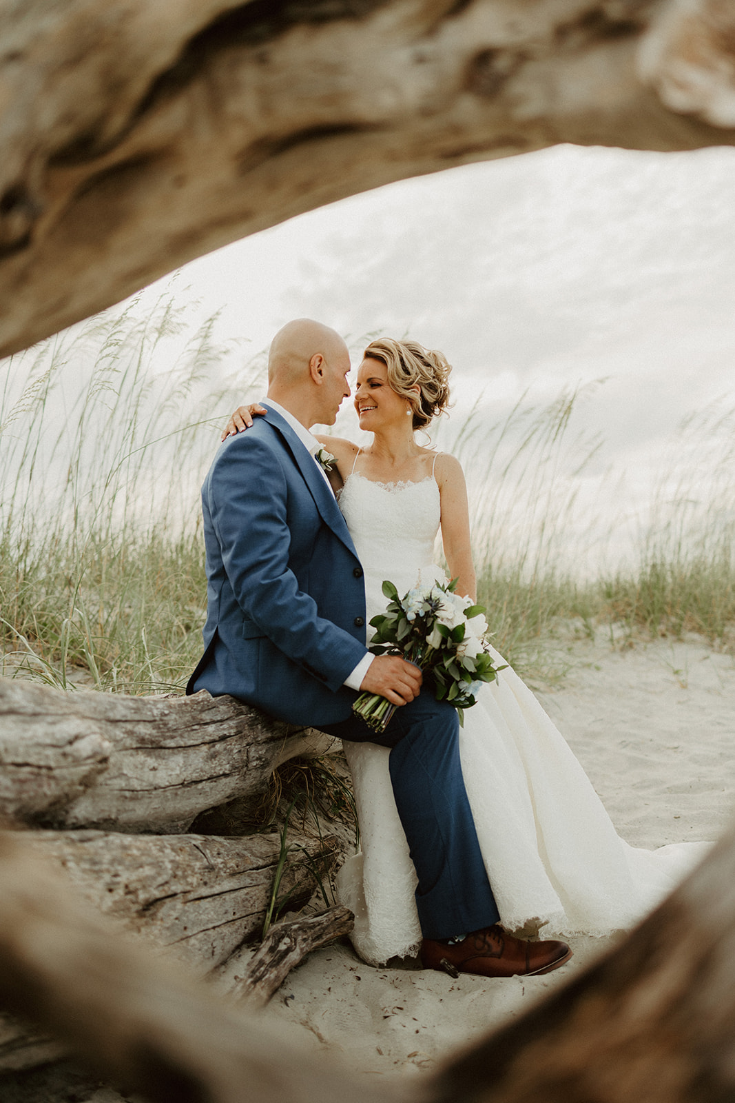 the bride and groom sitting on a log at the beach together