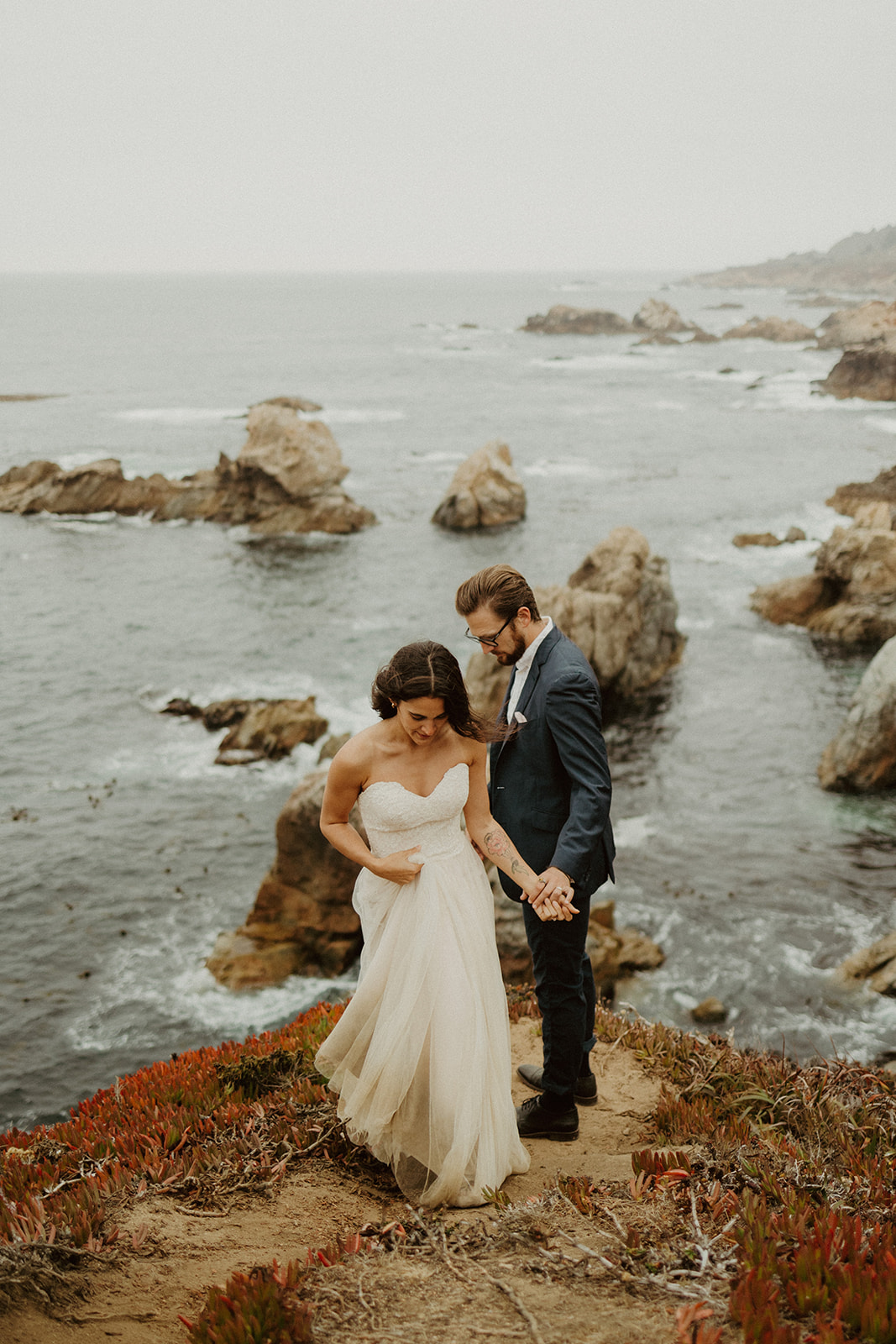 the wedding couple holding hands and walking away from the edge of the cliff at their foggy elopement in Big Sur