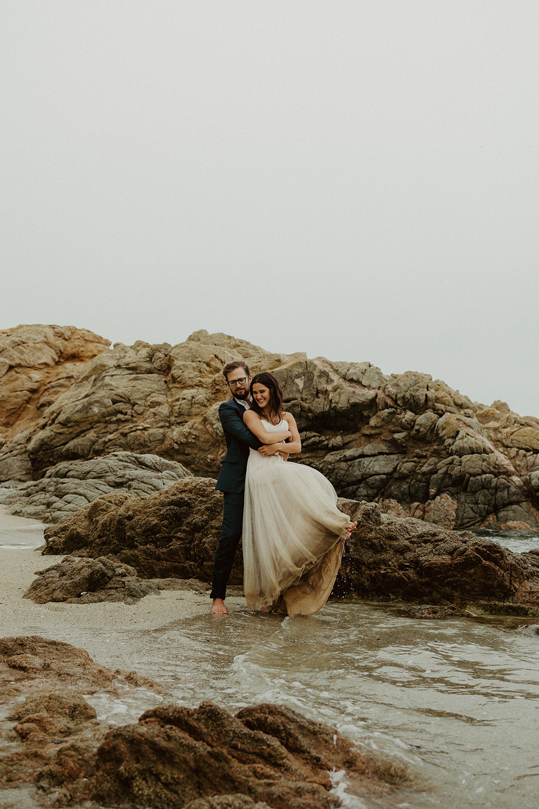 the groom lifting up the bride and twirling her around in the water at their foggy elopement in Big Sur